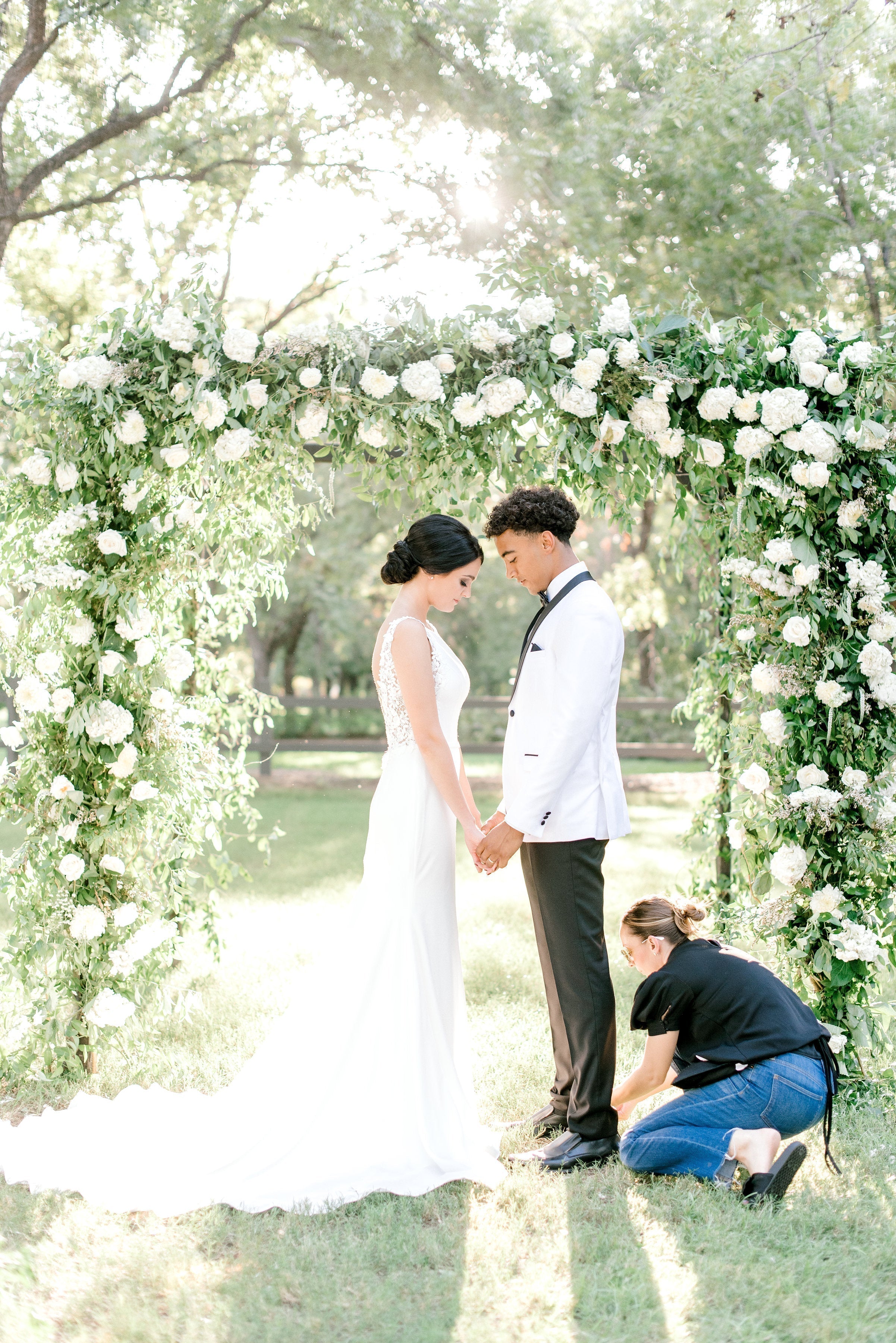 Bride and groom holding hands under a floral arch while a stylist adjusts the bride’s dress during an outdoor wedding ceremony.