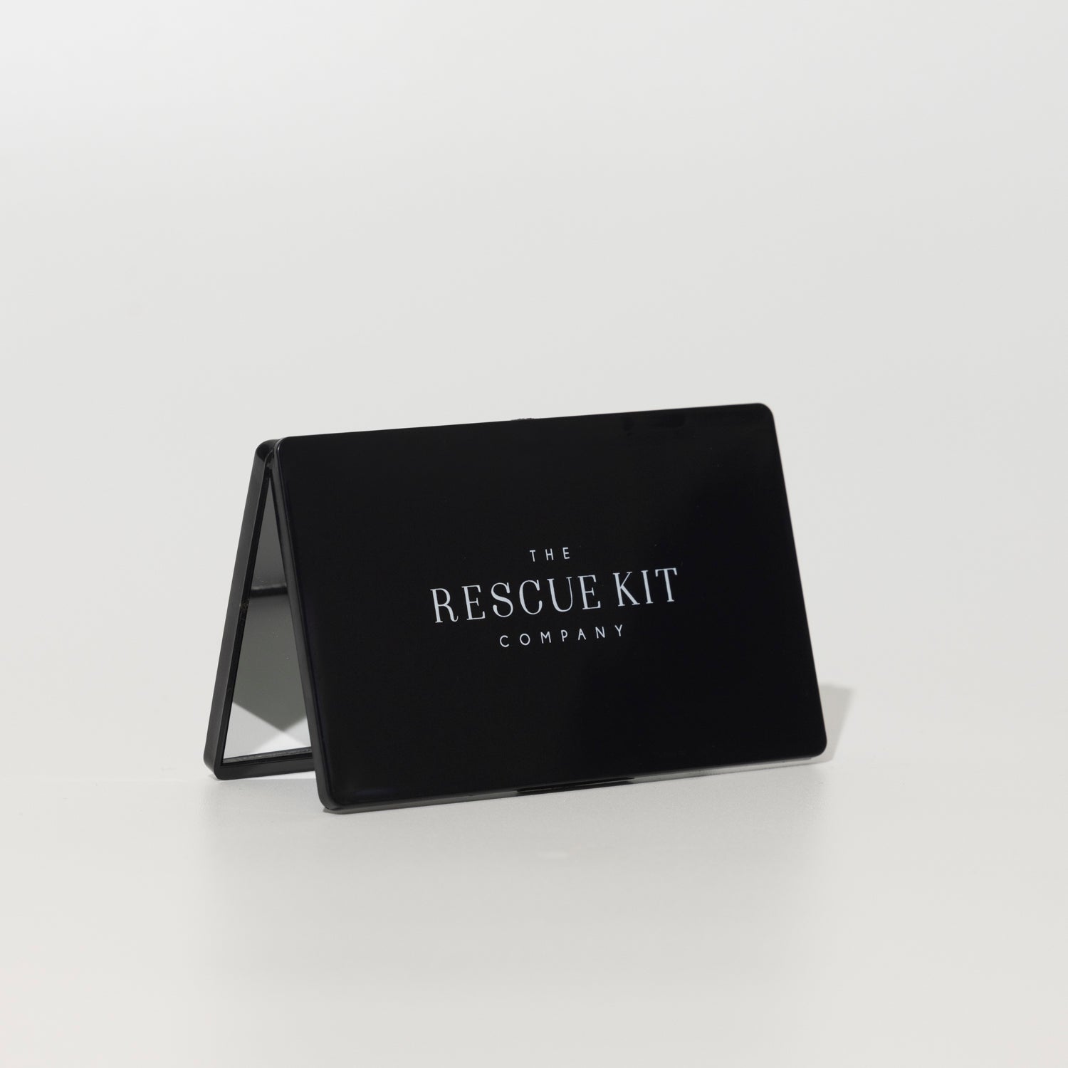 Black foldable vanity mirror with The Rescue Kit Company logo displayed on the front, propped open to reveal its compact, travel-friendly design.