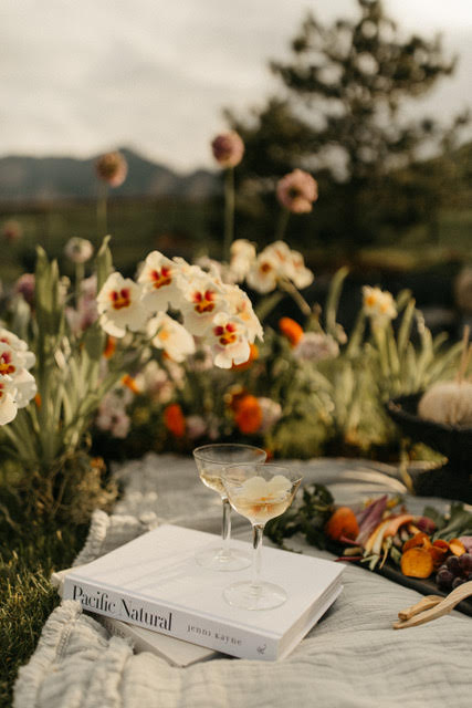 Elegant picnic scene with champagne coupes, a copy of Pacific Natural, and fresh vegetables on a blanket surrounded by blooming flowers in a garden setting.