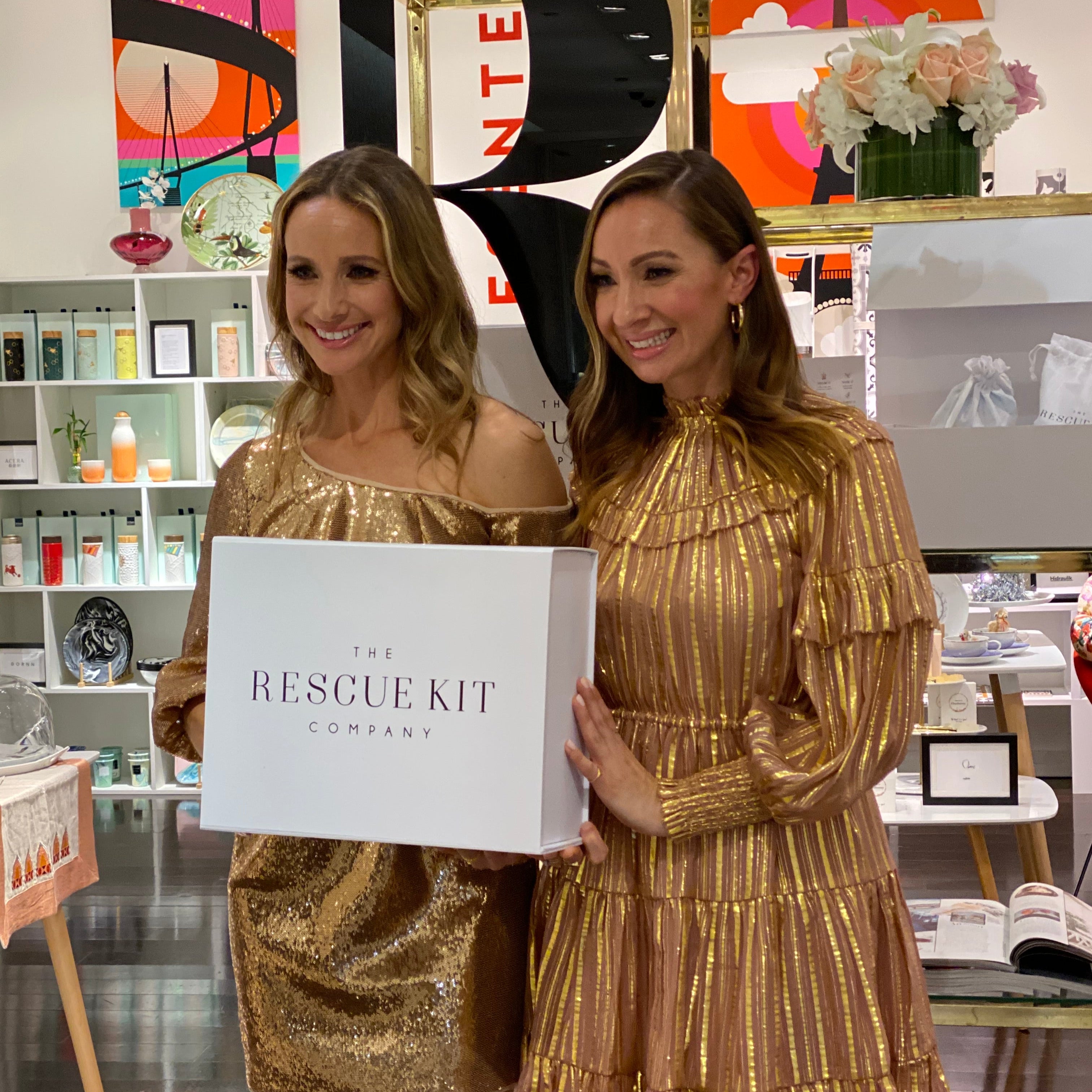 Founders of The Rescue Kit Company, Risa and Jess, at the launch of their company at a store within store concept in Saks 5th Avenue. Both sisters wear gold dresses, holding a white box with black company logo, standing in front of white shelving with beauty products and flowers.