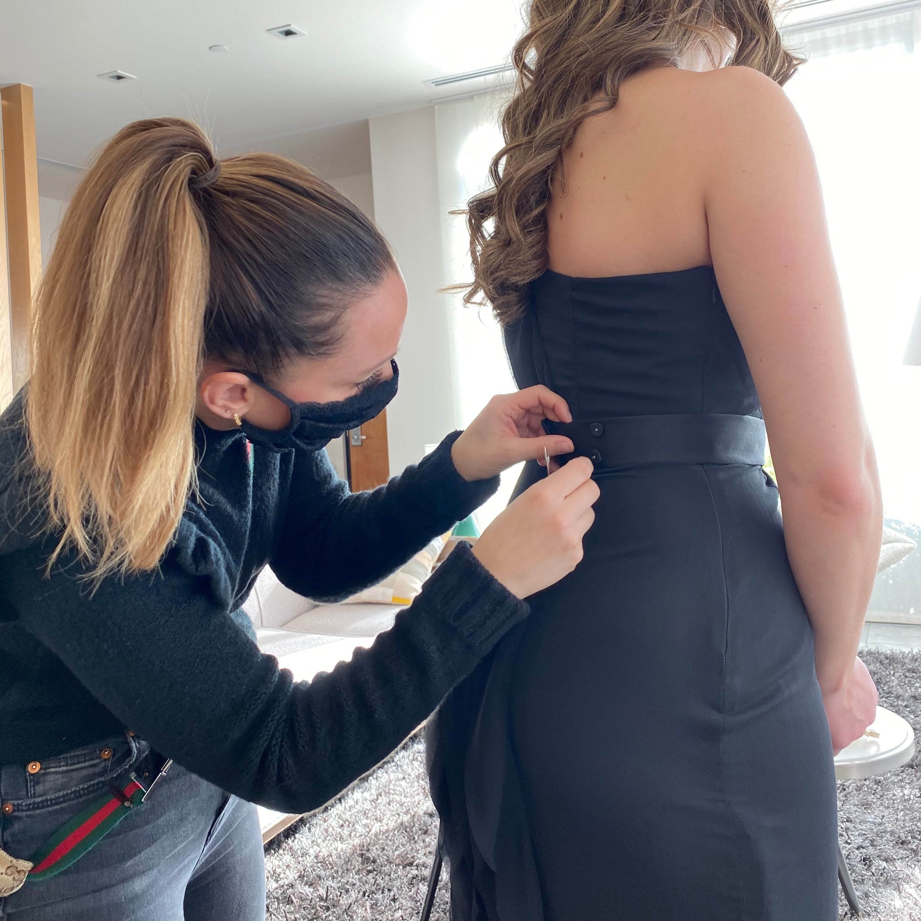 Stylist Risa Kostis, styling a bridesmaid during COVID. Risa wears a black mask and is sewing the back of the bridesmaid's black dress.